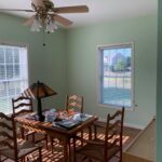 Freshly painted dining room with light green walls, featuring wooden furniture, a ceiling fan with light fixtures, and natural sunlight streaming through the windows.