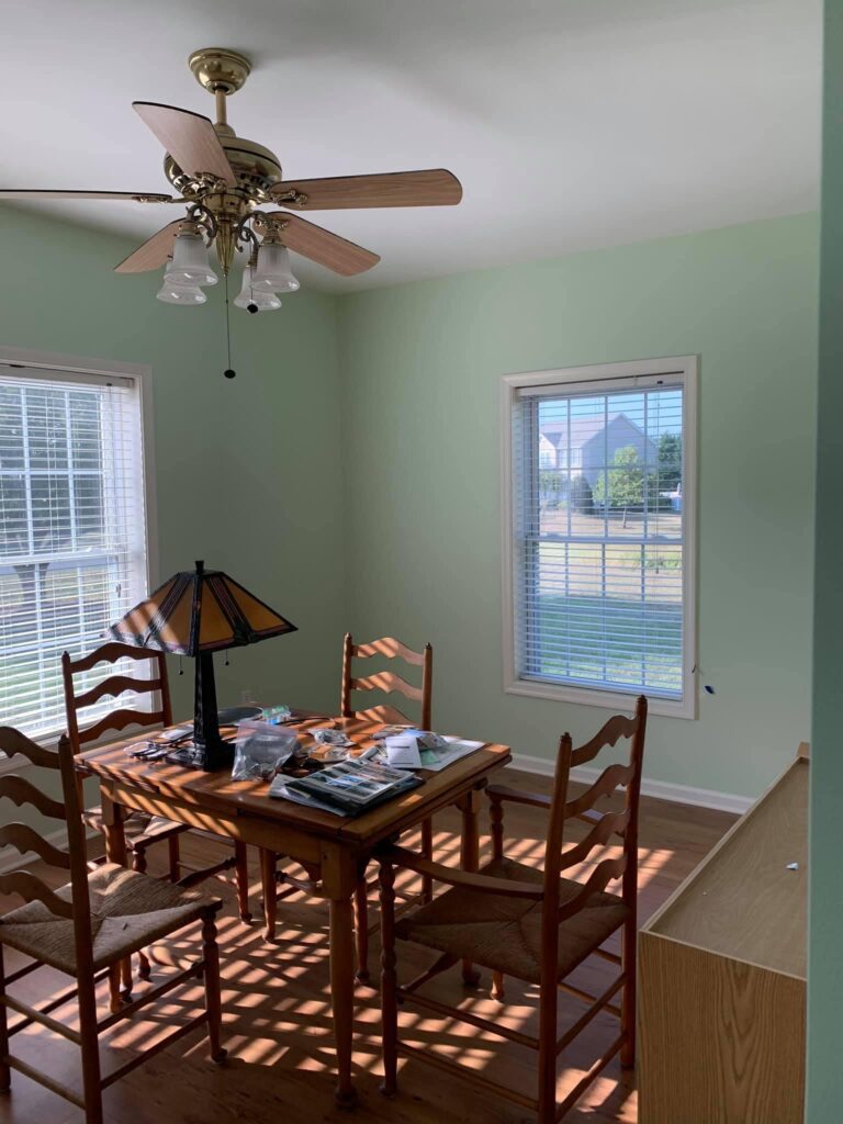 Freshly painted dining room with light green walls, featuring wooden furniture, a ceiling fan with light fixtures, and natural sunlight streaming through the windows.